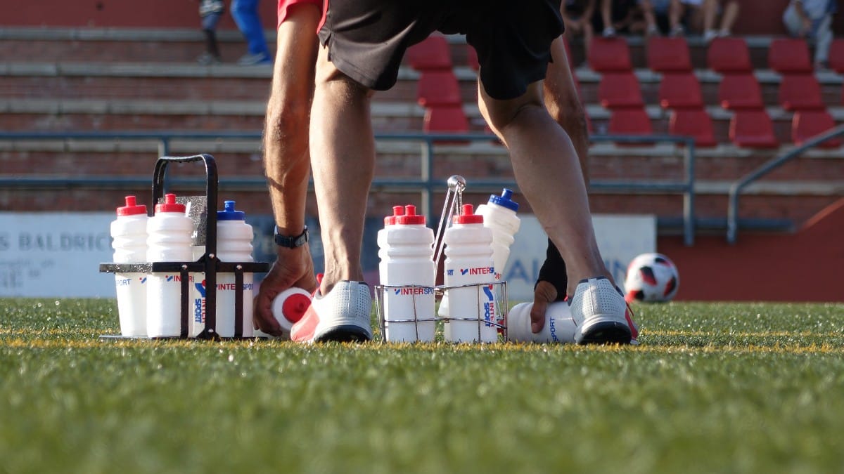 Un membre d’un cos tècnic recull ampolles d’aigua sobre la gespa d’un camp de futbol